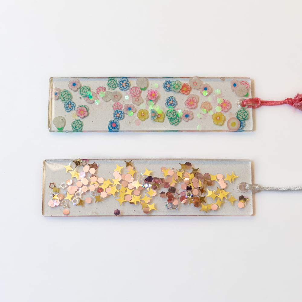 Two clear resin bookmarks, one with small flowers and the other with refective foil shapes that include stars and circles.
