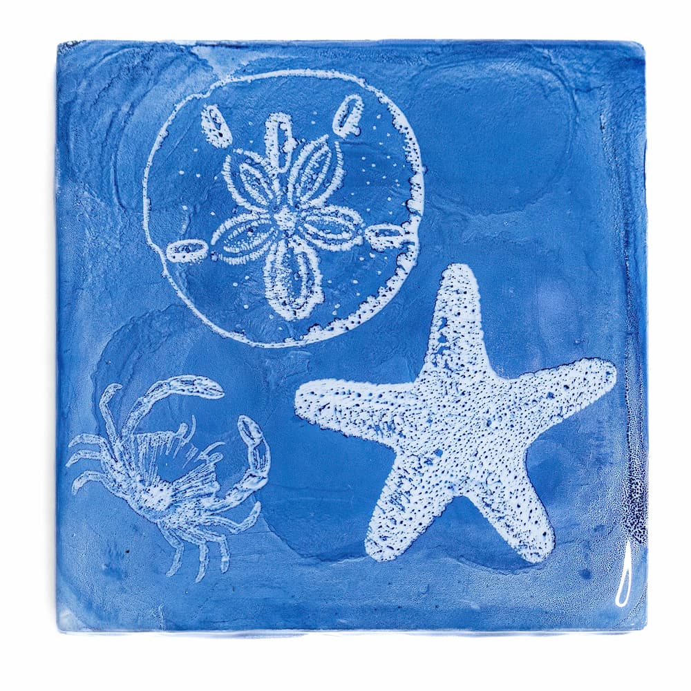 Blue alcohol ink resin coaster with white stamped images of sand dollar, crab, and starfish.